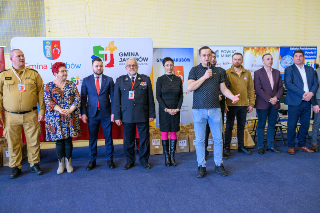 Grupa osób stojących w rzędzie na tle banerów gminnych. Mężczyzna po prawej stronie mówi do mikrofonu. Podłoga jest niebieska, a tło jasne.