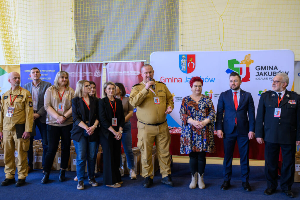 Grupa osób stoi w rzędzie w hali sportowej. Są ubrani formalnie, niektórzy w mundury. Tło jest żółto-niebieskie z banerami i logo Gmina Jakubów.