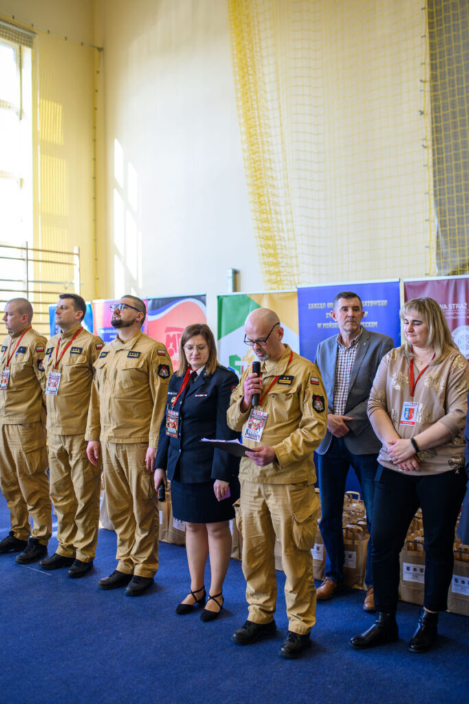 Grupa ludzi w mundurach stoi na niebieskim dywanie. W tle widoczne są banery z kolorowymi logo. Mężczyzna z mikrofonem czyta z kartki.