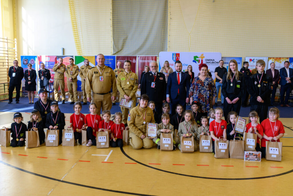 Grupa dzieci i dorosłych w mundurach stoi na tle banerów w hali sportowej. Dzieci mają medale i trzymają torby. Dominują kolory: czarny, czerwony i żółty.