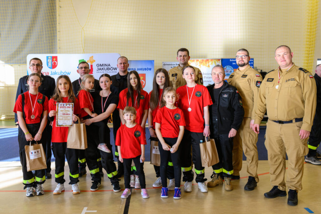 Grupa osób, głównie dzieci i młodzież, w czerwonych koszulkach, z medalami na szyi, stoi na tle baneru. Kolory dominujące to czerwony i beżowy. W tle widać napisy związane z gminą Jakubów.