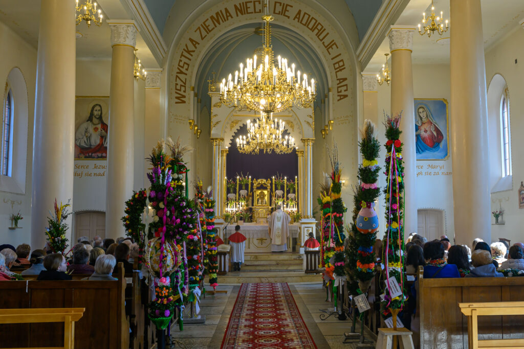 Wnętrze kościoła z jaskrawymi palmami wielkanocnymi. Jasne ściany i złoty żyrandol. Na ołtarzu świeci się wiele świec.