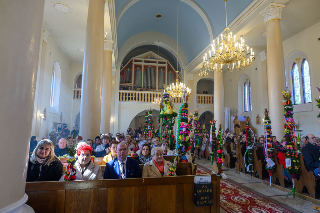 Wnętrze kościoła z ludźmi i kolorowymi palmami. Żyrandole na tle niebieskiego sklepienia. Jasne, ozdobne kolumny.