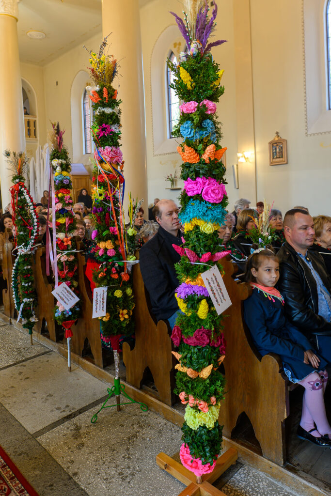 W jasnym wnętrzu kościoła widoczne są kolorowe palmy wielkanocne. Ozdobione kwiatami i wstążkami palmy stoją między ławkami. W tle zgromadzeni ludzie uczestniczą w mszy.