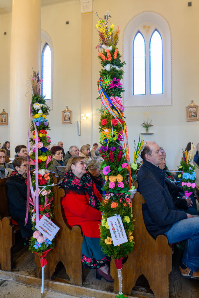 Kościół z osobami siedzącymi w ławkach, obok kolorowe palmy wielkanocne. Tło ma jasne ściany z dużymi oknami. Palmy ozdobione są kwiatami i wstążkami.