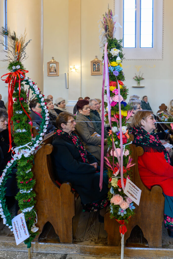 Wnętrze kościoła z ludźmi na ławkach, obok wysokie palmy wielkanocne z kolorowymi kwiatami i wstążkami. Ściany jasne, w tle witraże i obrazy.