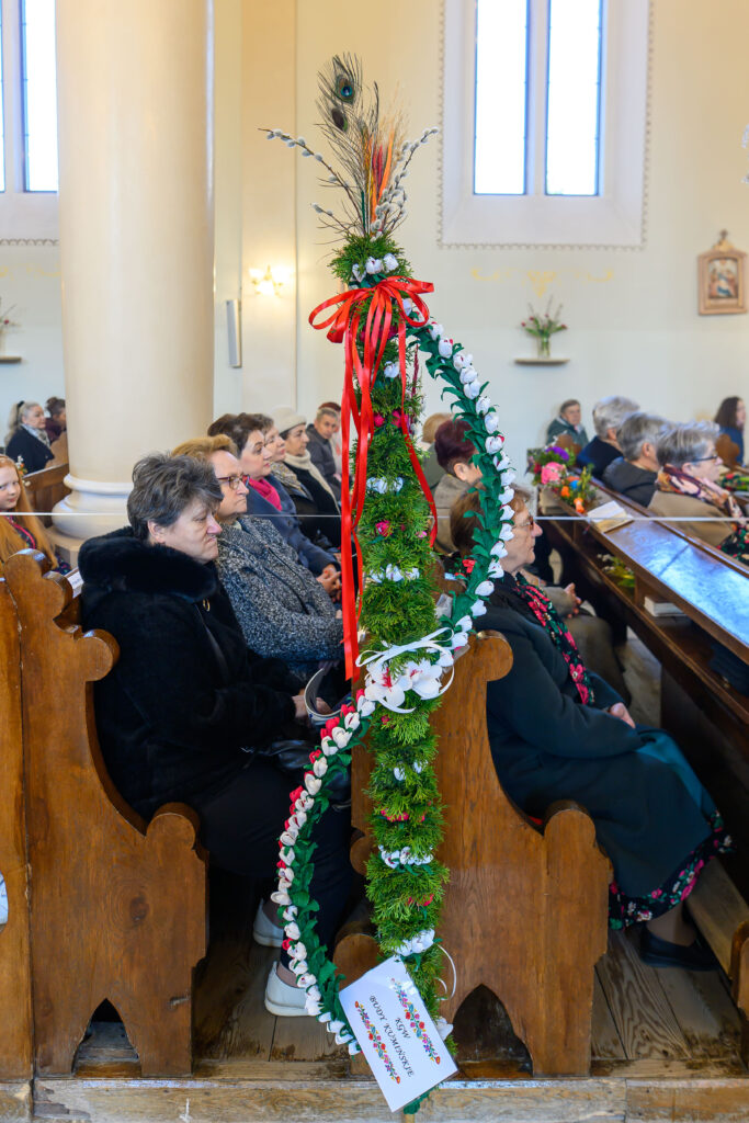 W kościelnej ławce siedzą ludzie, przed nimi przystrojona palma z barwnymi wstążkami. Dominują kolory zielony i czerwony. Tło to jasne wnętrze kościoła.