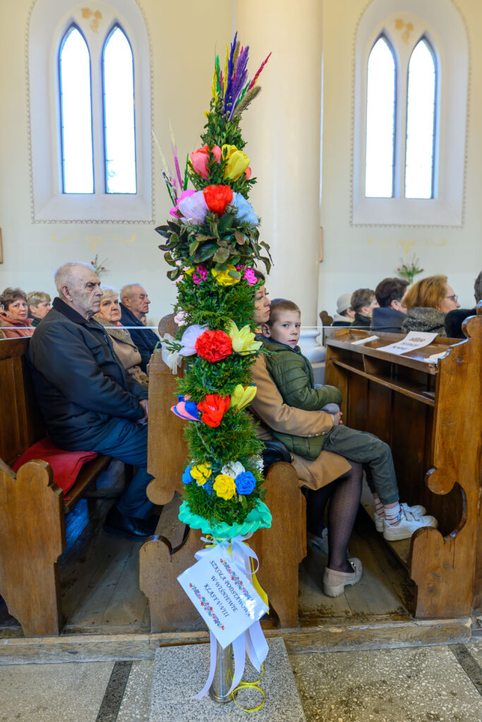 Kościół, wnętrze. Kolorowa palma wielkanocna w ławce, z boku siedzą ludzie. W tle witraże, atmosfera uroczysta.