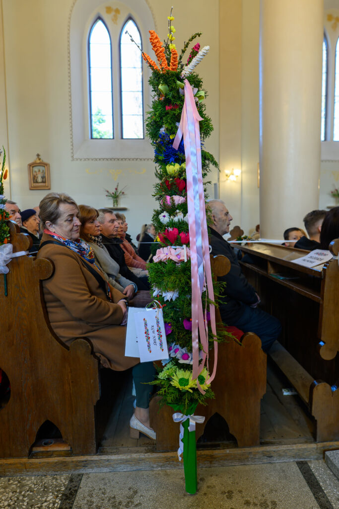Kościół w tle z wysoką, kolorową palmą wielkanocną ozdobioną kwiatami i wstążkami. Obok siedzą ludzie na ławkach. Wnętrze jasne, z dużymi oknami.
