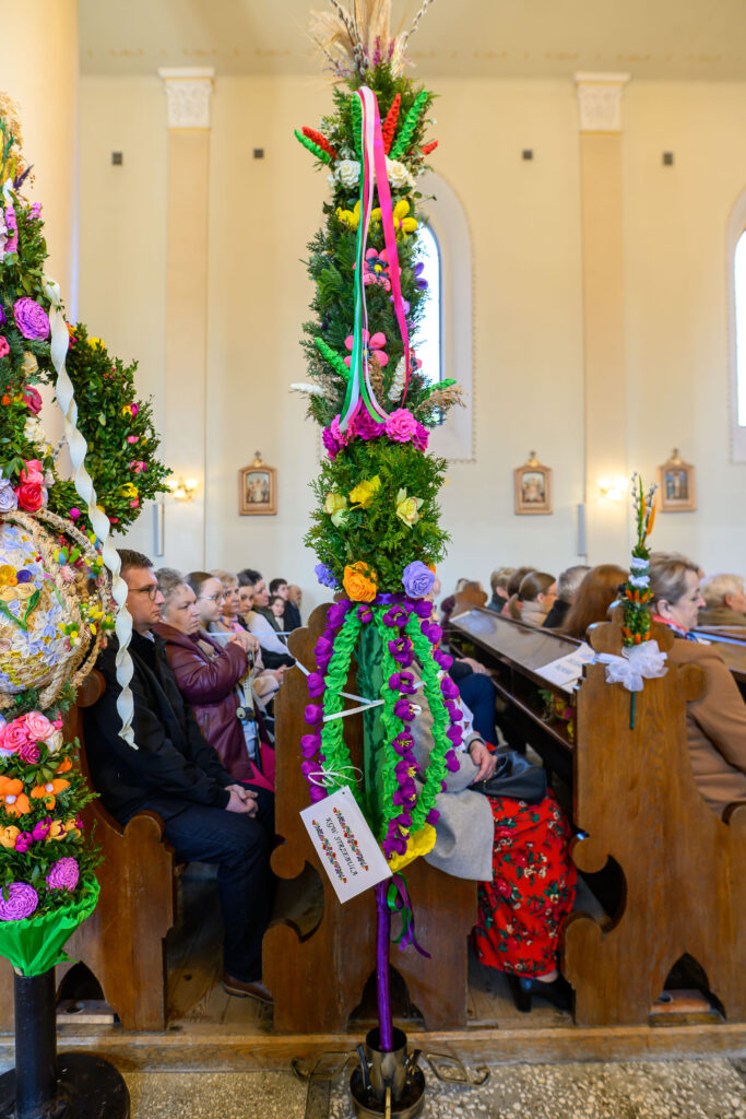 Kolorowa palma wielkanocna zdobi wnętrze kościoła. Barwne wstążki i kwiaty wyróżniają się na tle jasnych ścian i ławek. Uczestnicy mszy siedzą wewnątrz budynku.