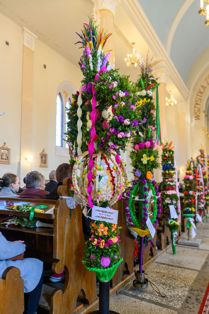 Kolorowe palmy wielkanocne stoją w drewnianych ławkach kościelnych. Dominują kolory zielony, fioletowy i różowy. Tło to wnętrze kościoła z jasnymi ścianami.