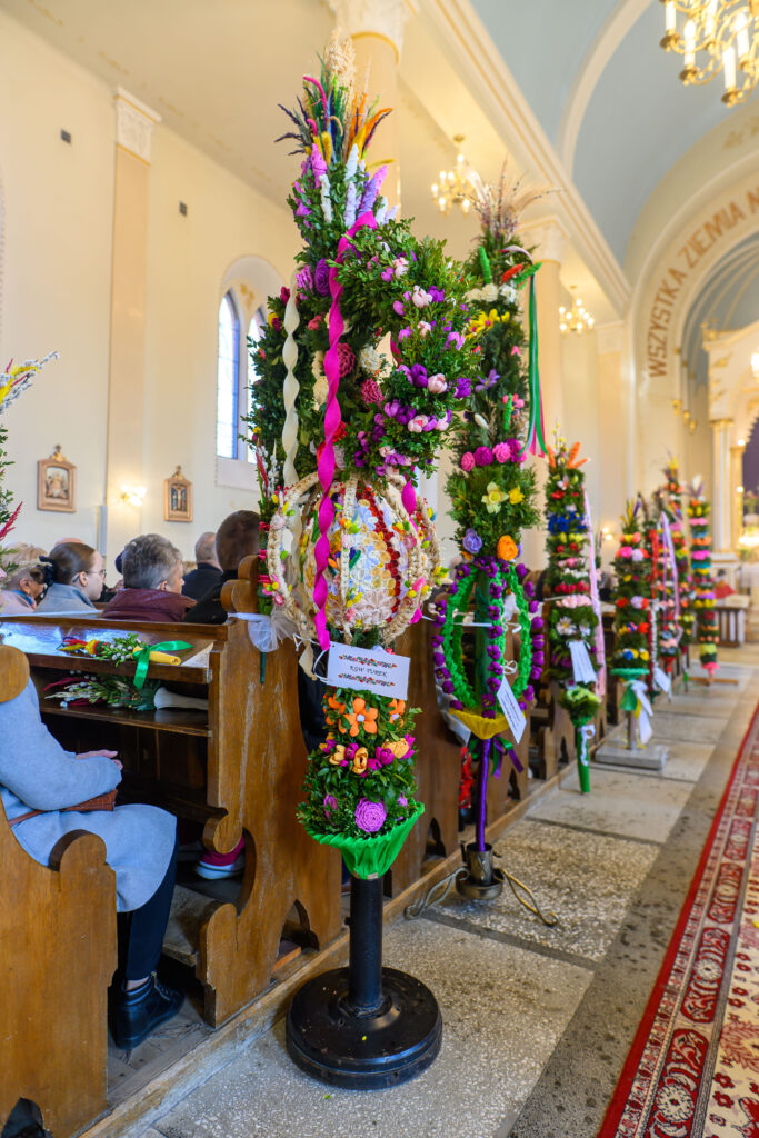 Wnętrze kościoła z wysokimi, kolorowymi palmami wielkanocnymi. Uczestnicy siedzą w drewnianych ławkach. Jasne ściany, złote żyrandole.