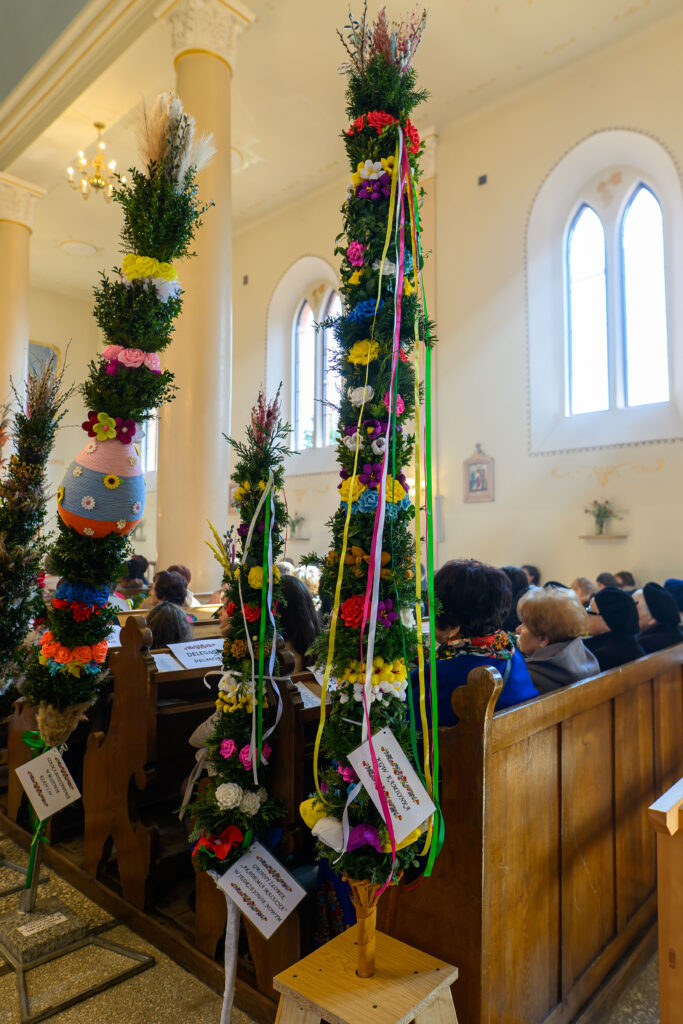Wnętrze kościoła z ozdobionymi palmami wielkanocnymi w żywych kolorach. Zawieszki z kolorowymi wstążkami. W tle ławki z ludźmi.