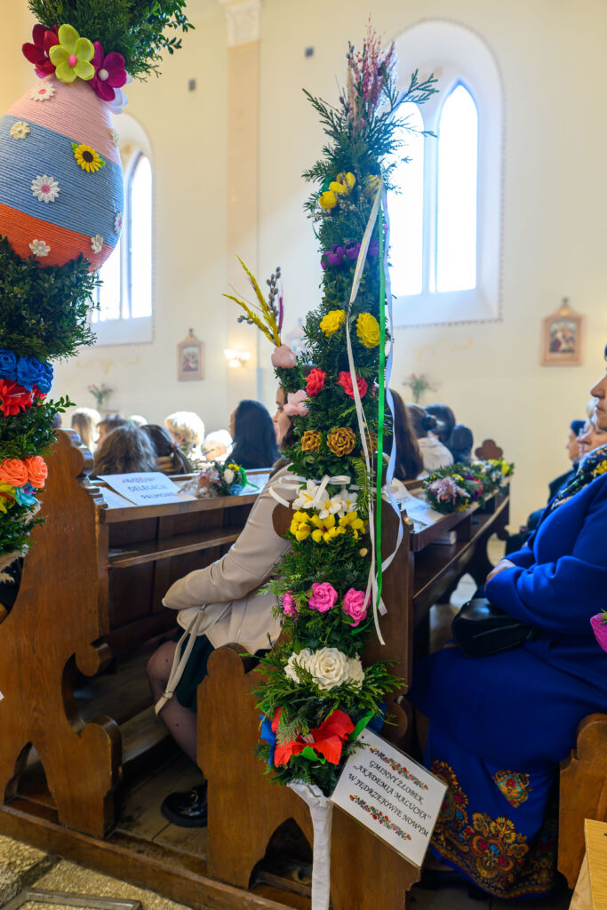 Kościół wypełniony ludźmi, w ławach ozdobne, kolorowe palmy z kwiatami i wstążkami. Jasne wnętrze, drewniane ławki. Atmosfera świąteczna.