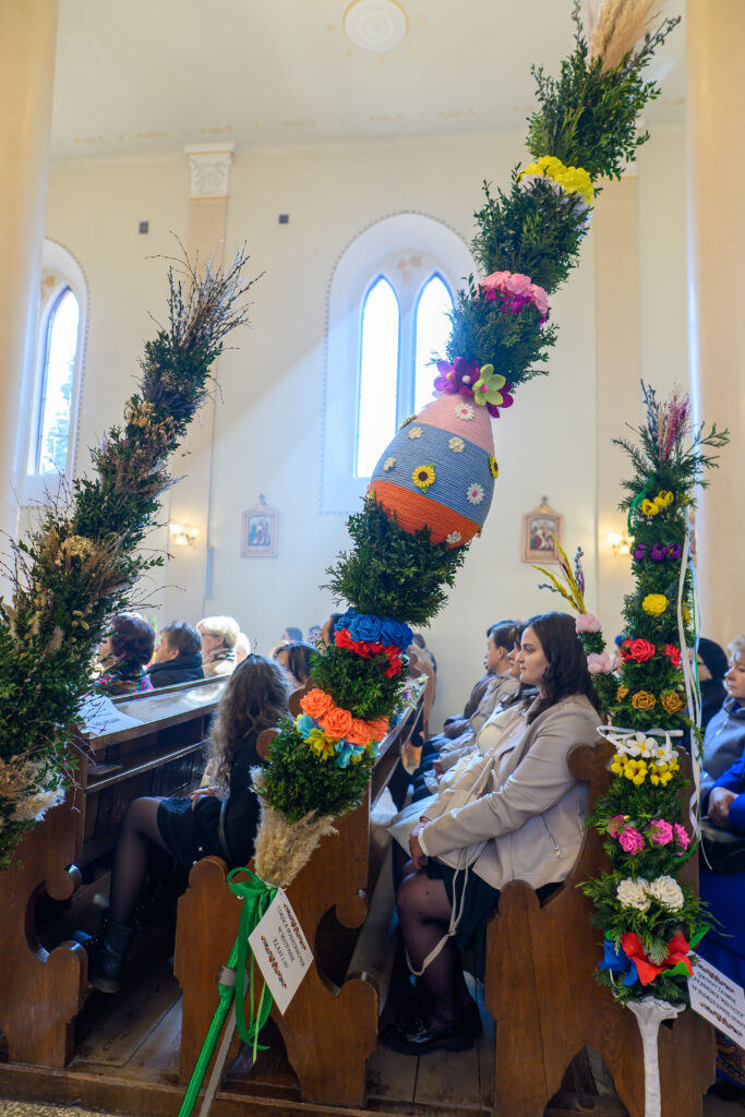 Wnętrze kościoła z ludźmi trzymającymi palmy wielkanocne. Palmy są udekorowane kolorowymi kwiatami i wstążkami. W tle jasne ściany i okna łukowe.