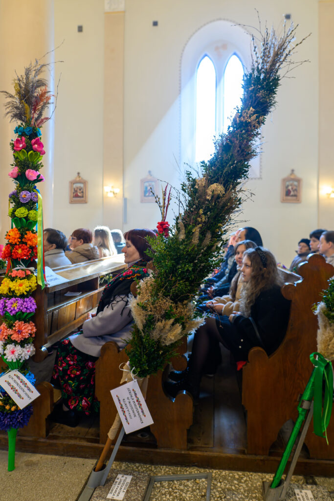 Sala kościelna z kolorowymi palmami wielkanocnymi. Ludzie siedzą na drewnianych ławach, światło wpada przez okno. Ściany jasne, na nich obrazy.