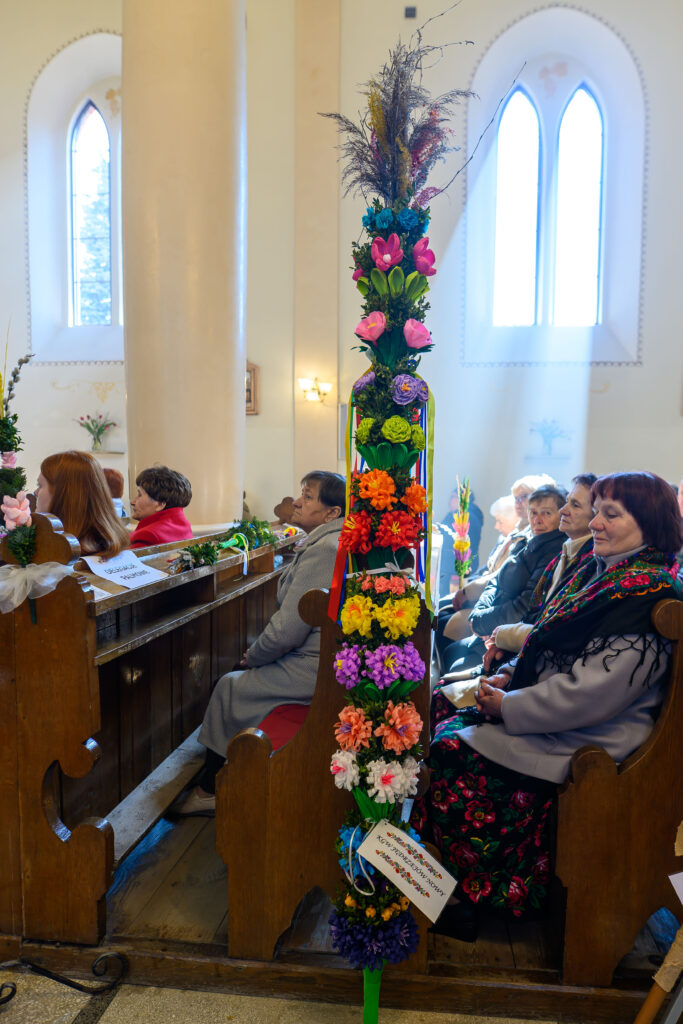 Wnętrze kościoła, ludzie siedzą w drewnianych ławkach. Przy jednej z ławek stoi wysoka, kolorowa palma wielkanocna z kwiatami. Przez okna wpadają promienie słoneczne, tworząc jasne tło.