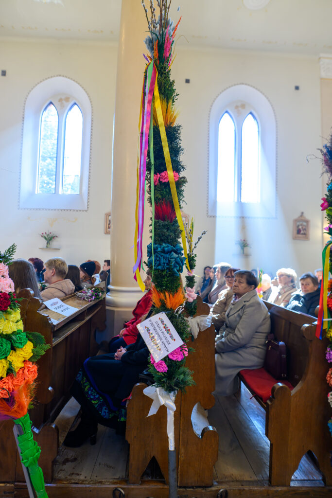 W kościele ludzie siedzą na drewnianych ławkach, trzymając barwne palmy wielkanocne. Palmy zdobią kolorowe kwiaty i wstążki. Światło wpada przez wysokie, łukowate okna.