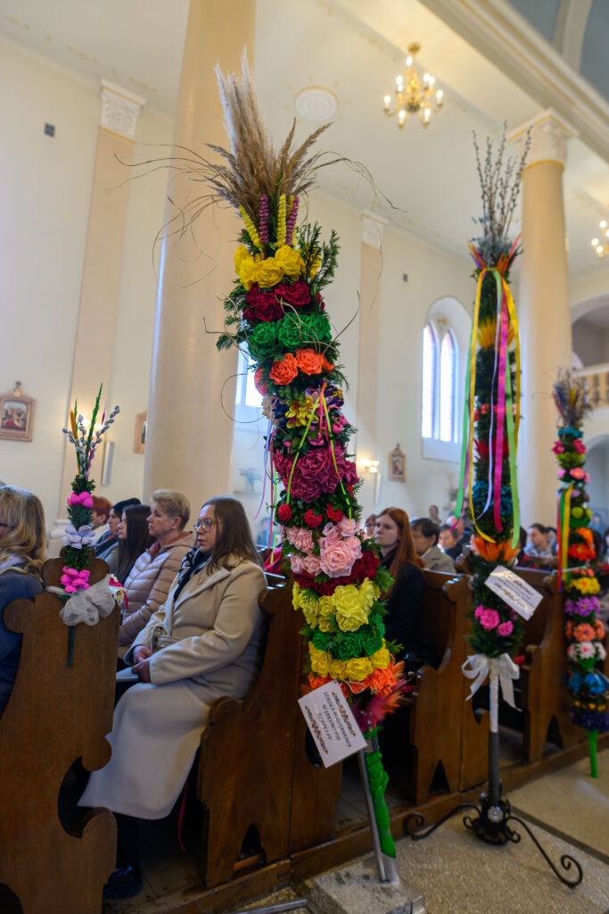 Wnętrze kościoła z wysokimi, kolorowymi palmami wielkanocnymi z kwiatów w intensywnych barwach. W tle widać jasne ściany i żyrandole. Osoby siedzą na drewnianych ławkach.