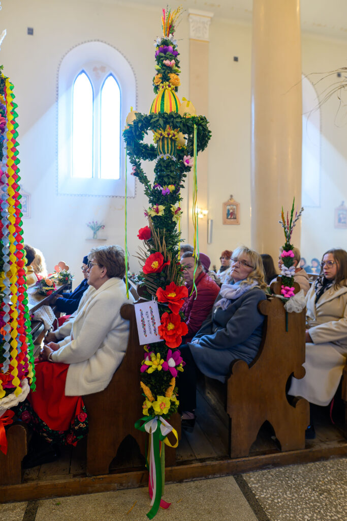 Kościół z ludźmi siedzącymi w ławkach, ozdobionych wysokimi palmami wielkanocnymi. Jasne, ciepłe wnętrze z dużymi oknami. Kolorowe kwiaty i zielone liście dominują w dekoracjach.
