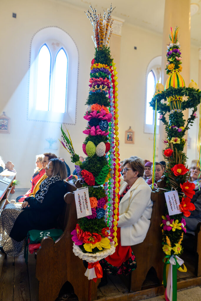 Kolorowe palmy wielkanocne z kwiatami i wstążkami w kościele. Ludzie siedzą w ławkach. Promienie słońca wpadają przez okna.