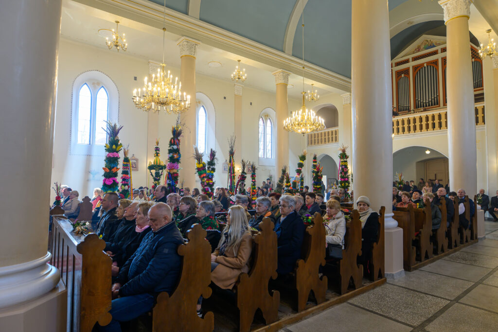 Ludzie siedzą w ławkach w kościele, trzymając kolorowe palmy. Jasne wnętrze z dużymi żyrandolami i tego samego koloru kolumnami.