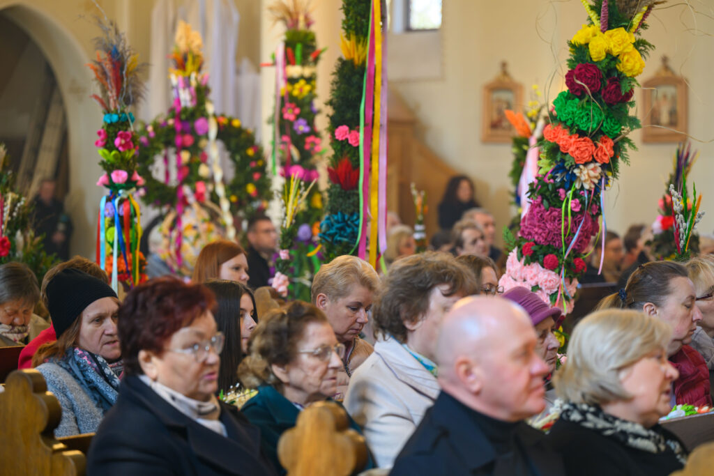 Grupa ludzi siedzi w kościele, otoczona kolorowymi palmami wielkanocnymi. Dominują barwy różowe, zielone i żółte. Na tle widać jasne wnętrze świątyni.