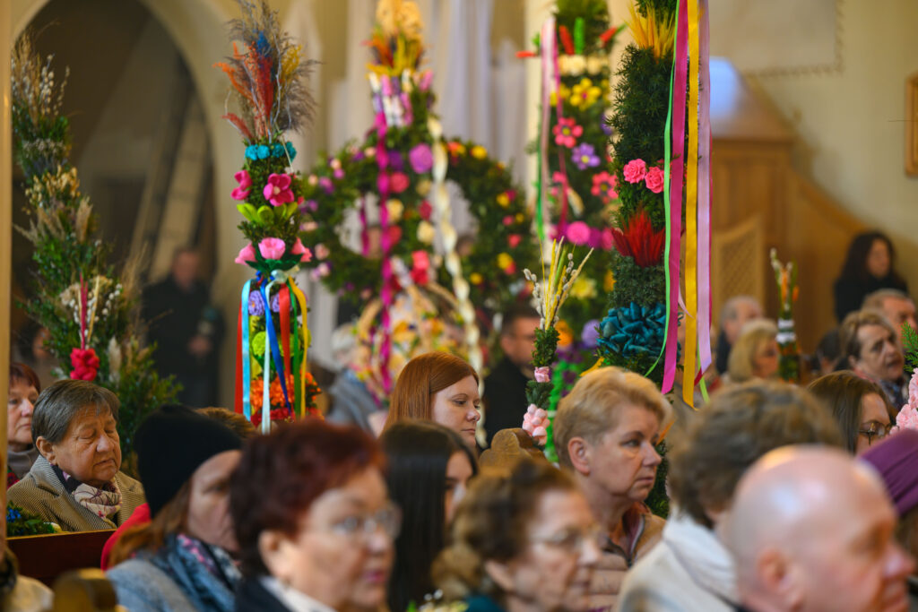 Na obrazie widać wiernych w kościele, trzymających kolorowe palmy wielkanocne. Dominuje zieleń, róż i żółć. Tło stanowi jasne wnętrze świątyni.