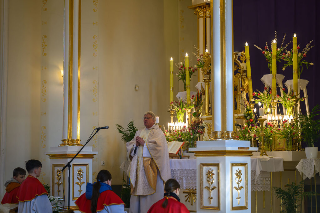 Kapłan w białej szacie stoi przy ołtarzu z kwiatami i świecami. W tle złote kolumny i bogata dekoracja. Ministranci w czerwonych pelerynach klęczą.