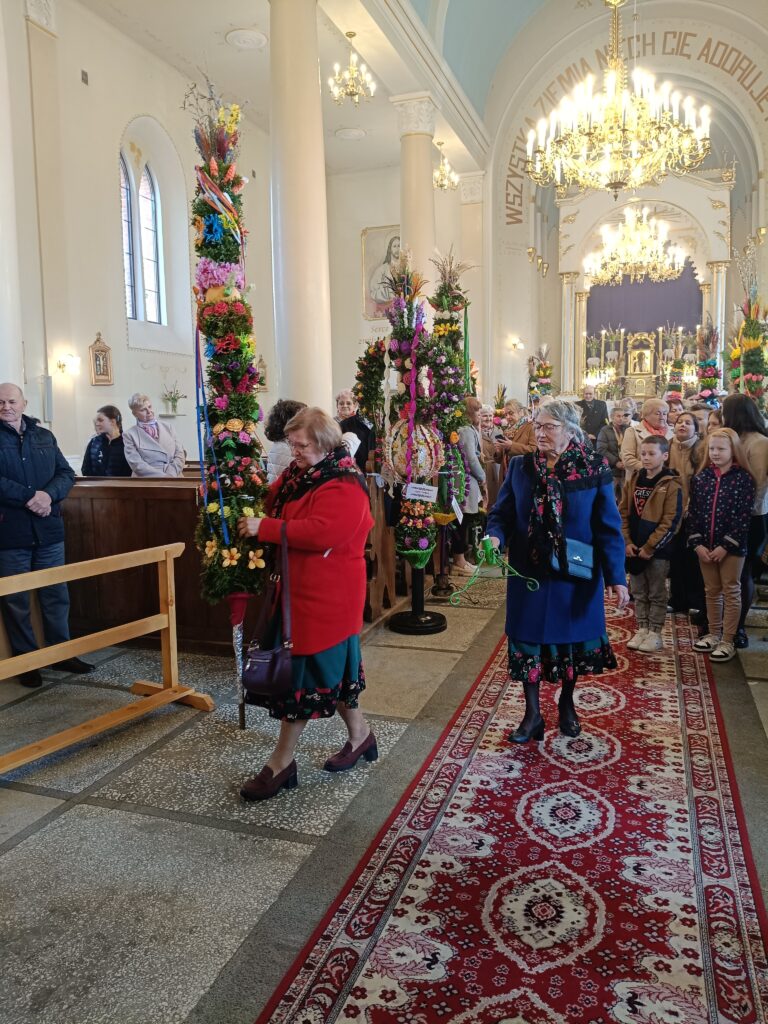 Wnętrze kościoła z ludźmi trzymającymi ozdobne palmy. Dominują jasne kolory, w tle widać żyrandol. Płytki podłogowe i dekoracyjny dywan prowadzą przez nawę.