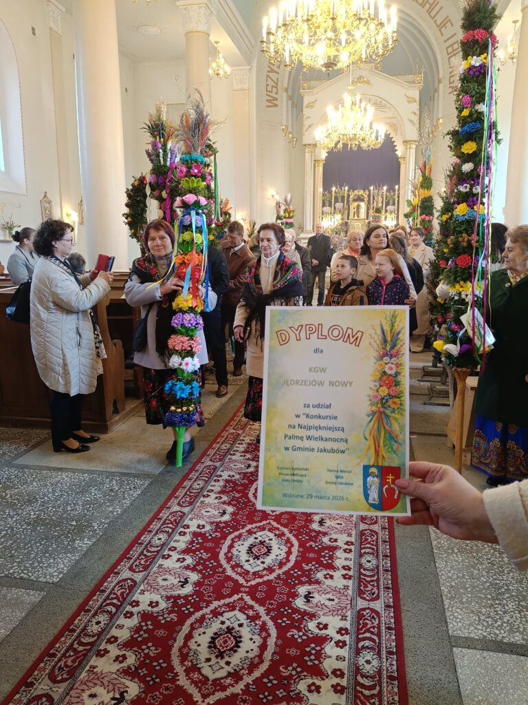 Grupa osób w kościele z kolorowymi palmami i dyplomem. Jasne wnętrze zdobią żyrandole, na podłodze czerwony dywan. Tło jest bogato zdobione, z kolumnami i ołtarzem.