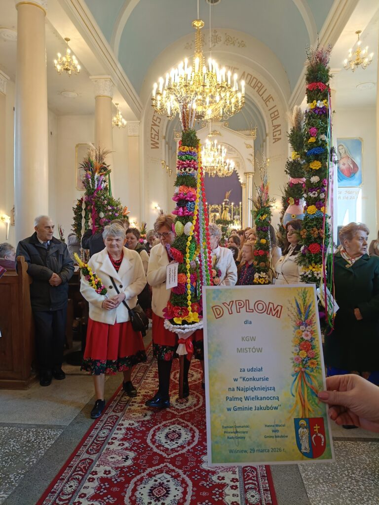 Wnętrze kościoła z ludźmi trzymającymi kolorowe palmy wielkanocne. Na pierwszym planie widoczny jest dyplom. Tło ozdobione żyrandolami i malowidłami.