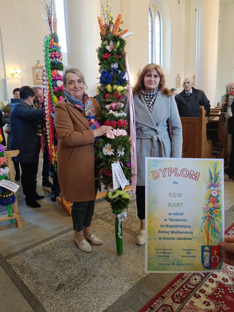 Dwie kobiety trzymają kolorowe palmy w kościele. Tło jest jasne z wysokimi oknami. Widać dyplom za konkurs palm wielkanocnych.