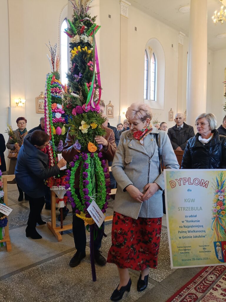 Na zdjęciu widoczne są osoby w kościele z dużą, kolorową palmą wielkanocną. Palma jest udekorowana kwiatami i wstążkami w intensywnych kolorach, takich jak fiolet, żółty i zielony. W tle widać jasne ściany i oz