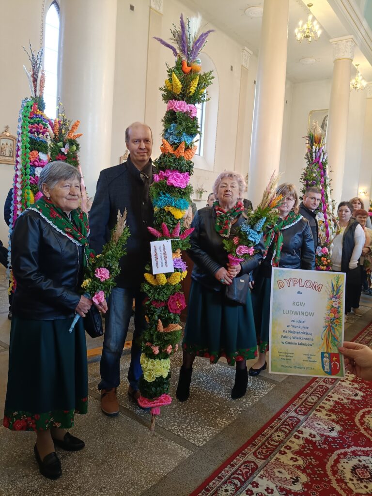Grupa ludzi trzyma wysokie, kolorowe palmy z kwiatami w kościelnym wnętrzu. Tło jest jasne z białymi filarami. Jedna osoba pokazuje dyplom z napisem.