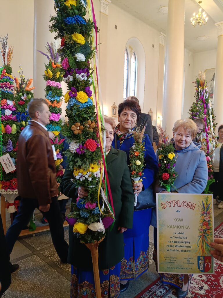 Kilka kobiet trzyma kolorowe palmy wielkanocne w kościele. W tle widoczne są jasne ściany i okna. Jedna osoba trzyma dyplom z napisem KGW KAMIONKA.