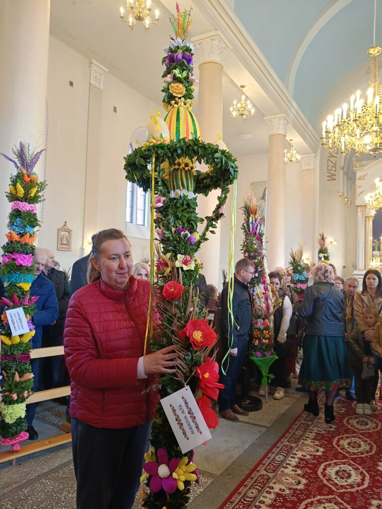 Zdjęcie przedstawia osobę trzymającą dużą, kolorową palemkę wielkanocną z kwiatami w kościele. Dominują kolory zielony, czerwony i żółty. W tle są ludzie i jasne wnętrze z kolumnami i żyrandolami.