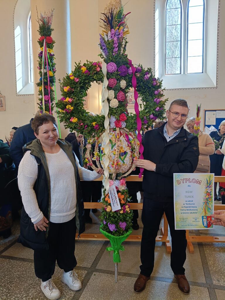 Osoby trzymają wysoką, kolorową palmę z kwiatami w kościele. Tło jasne, z oknami; w rękach dyplom. Dekoracje zielono-fioletowe.