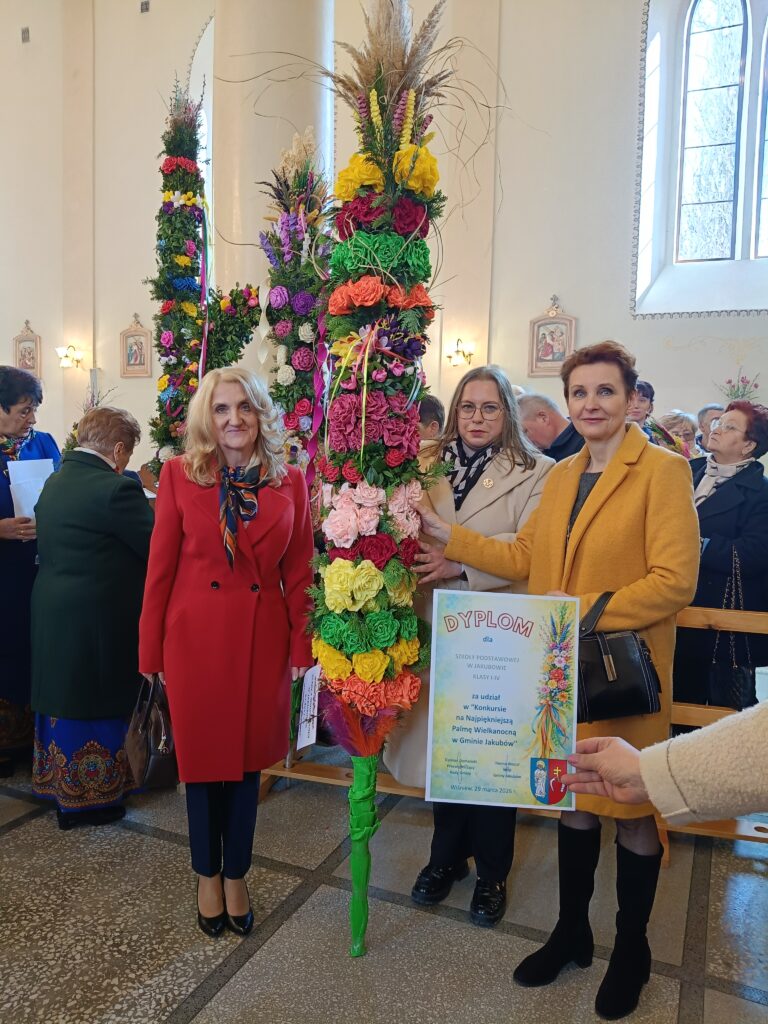 Trzy kobiety stoją z kolorowymi palmami wielkanocnymi w kościele. Tło jasne, wysokie okna, dekoracje kwiatowe. Jedna trzyma dyplom.