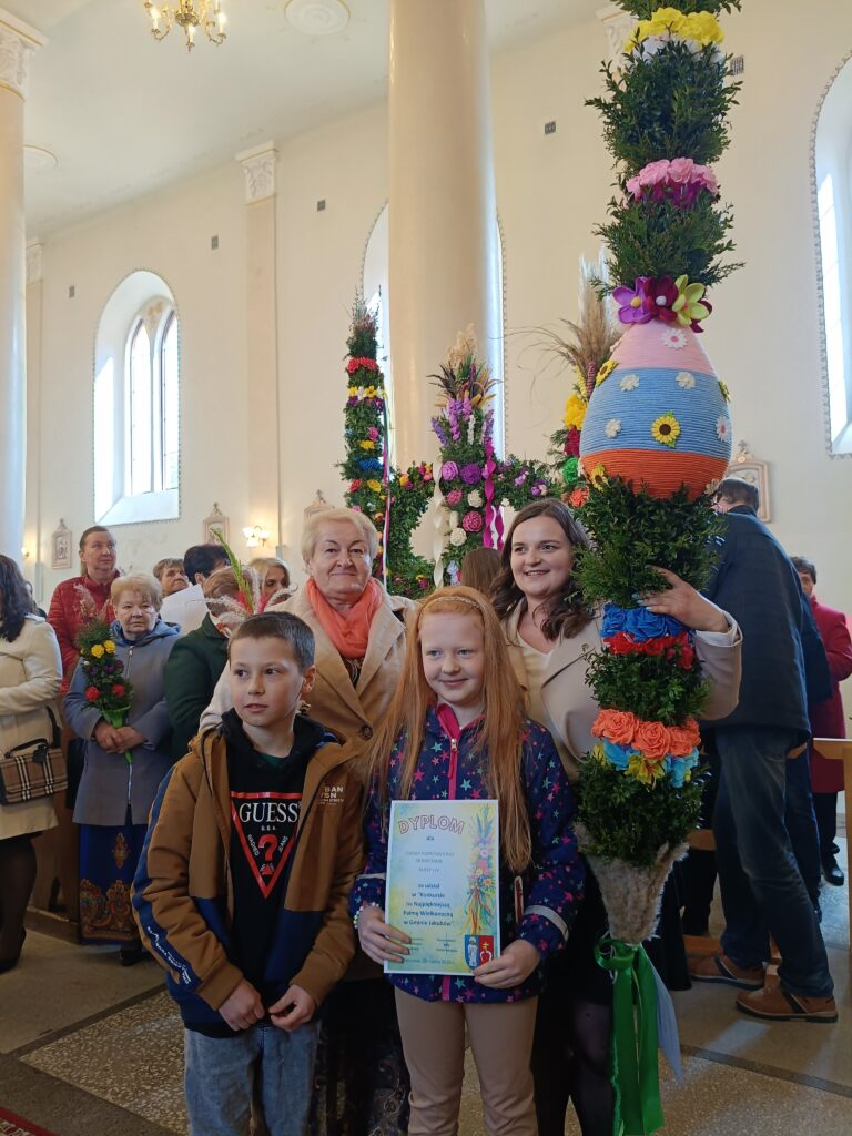 Grupa ludzi w kościele trzyma kolorowe palmy wielkanocne. Na zdjęciu są dzieci z dyplomem i starsze osoby. Wnętrze jasne, z dużymi oknami i kolumnami.