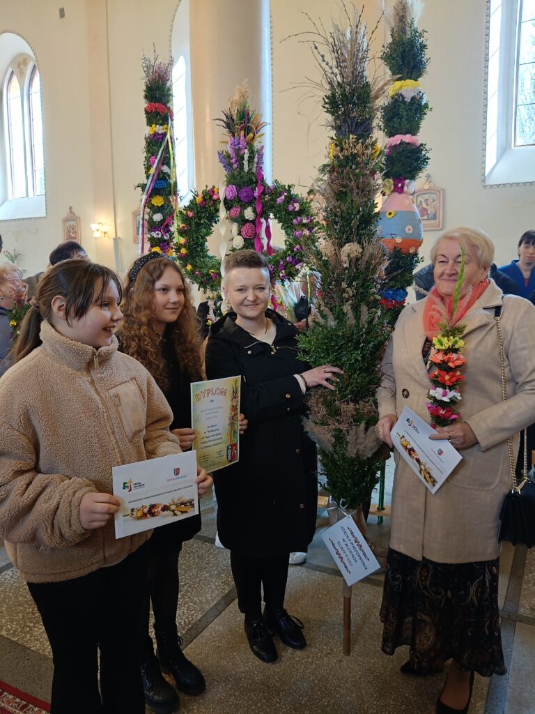 Grupa osób w jasnym wnętrzu, trzyma duże, kolorowe palmy wielkanocne. Palmy zdobione są kwiatami i wstążkami. W tle widać duże okna wpuszczające światło.