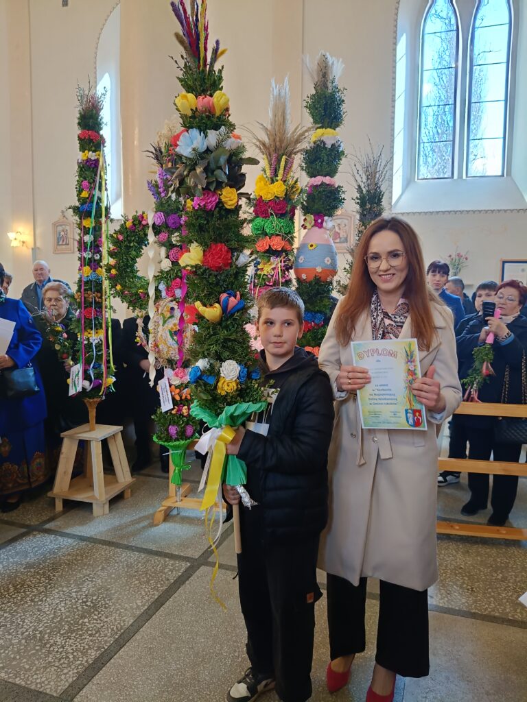 Na zdjęciu widać chłopca i kobietę w kościele, trzymających wielkanocne palmy. Palmy są kolorowe, ozdobione kwiatami i wstążkami. W tle znajdują się inni ludzie i duże okno.