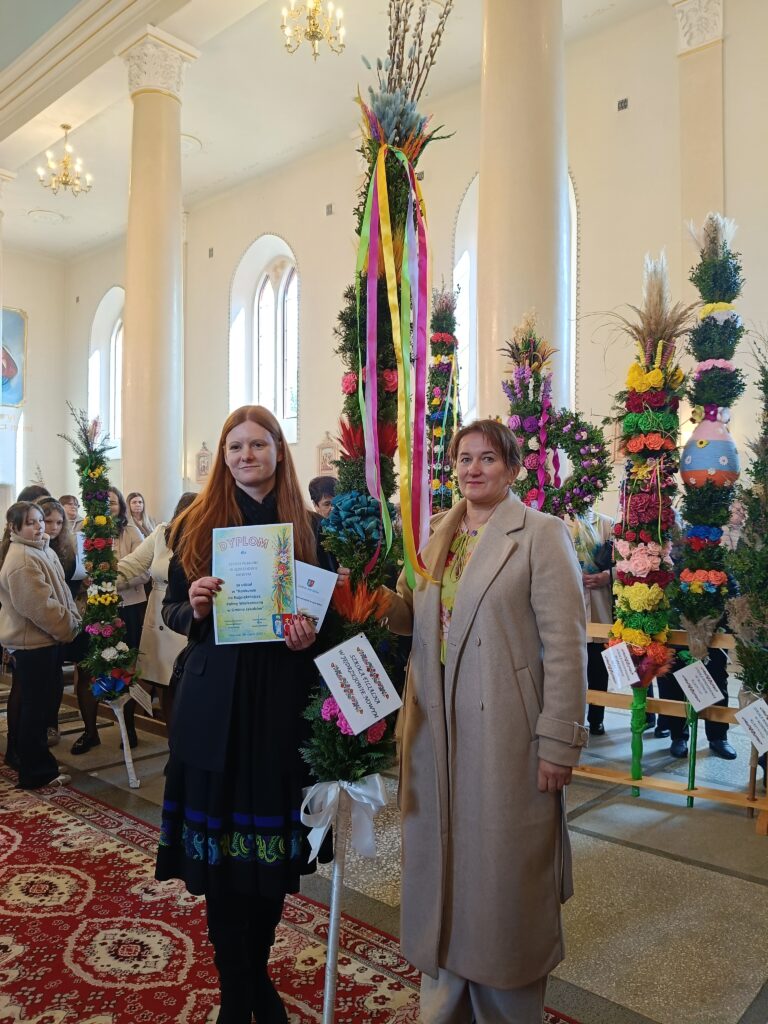 Na zdjęciu dwie kobiety z dyplomem stoją w kościele wśród wielkanocnych palm. Kolorowe palmy zdobią jasne wnętrze z białymi kolumnami. Uczestnicy w tle tworzą uroczystą atmosferę.