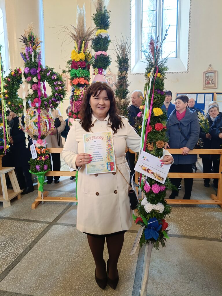Kobieta w jasnym płaszczu trzyma dyplom, stojąc obok kolorowych palm wielkanocnych. W tle widać ludzi w jasnym wnętrzu kościoła. Dominują kolory zielony i różne odcienie kwiatów.