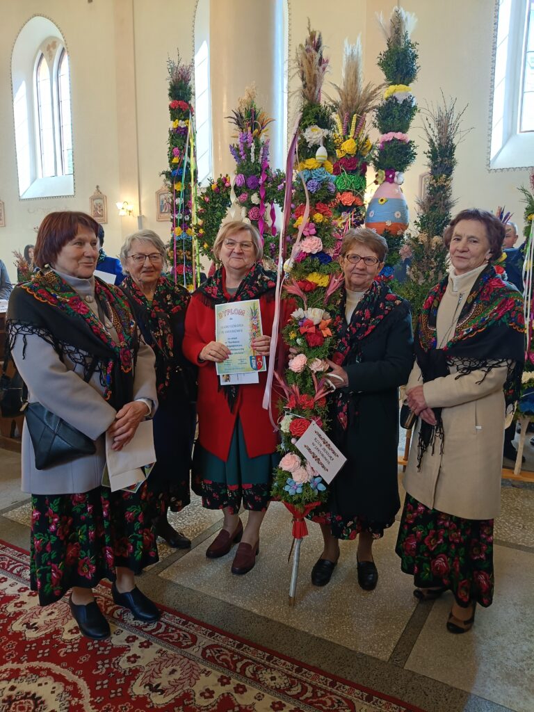 Pięć kobiet w strojach ludowych stoi w kościele z ozdobnymi palmami wielkanocnymi. Tło jest jasne z wysokimi oknami. Jedna kobieta trzyma dyplom.