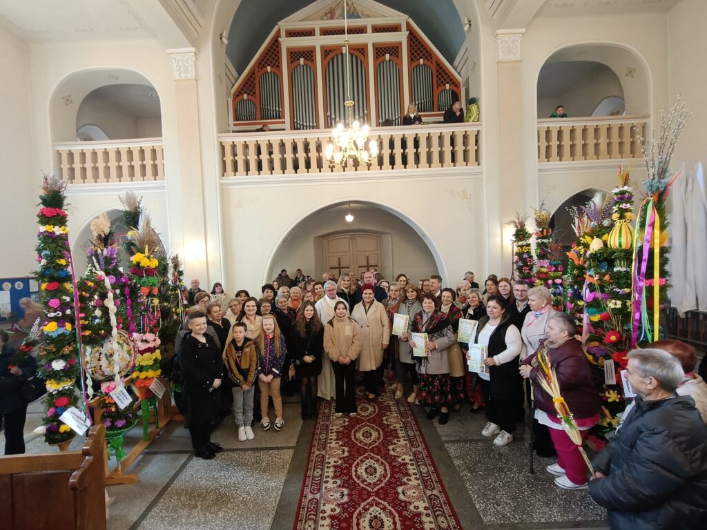 Grupa ludzi stoi w jasnym wnętrzu kościoła, ozdobionym kolorowymi palmami wielkanocnymi. Tło jest kremowe, z ozdobnym balkonem i organami. W centrum na dywanie znajduje się zgromadzenie przestrzegające tradycji.