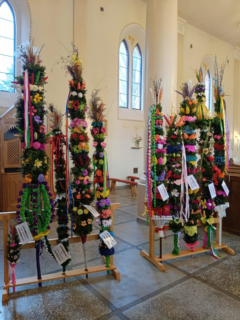 Kolorowe palmy wielkanocne stoją w jasnym wnętrzu kościoła. Przyozdobione są kwiatami i wstążkami w różnych kolorach. W tle widać okna z łukowatymi witrażami.