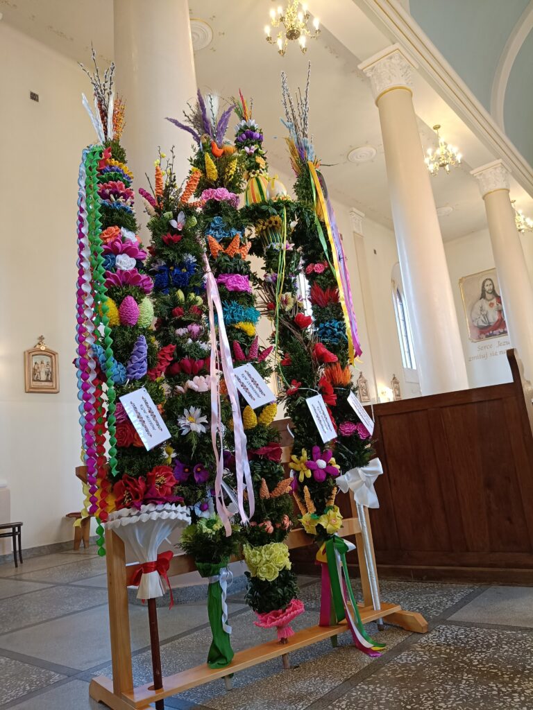 Na zdjęciu widoczne są kolorowe palmy wielkanocne z kwiatami i wstążkami. Tło stanowi jasne wnętrze kościoła z kolumnami. Dekoracje ozdobione są barwnymi elementami.