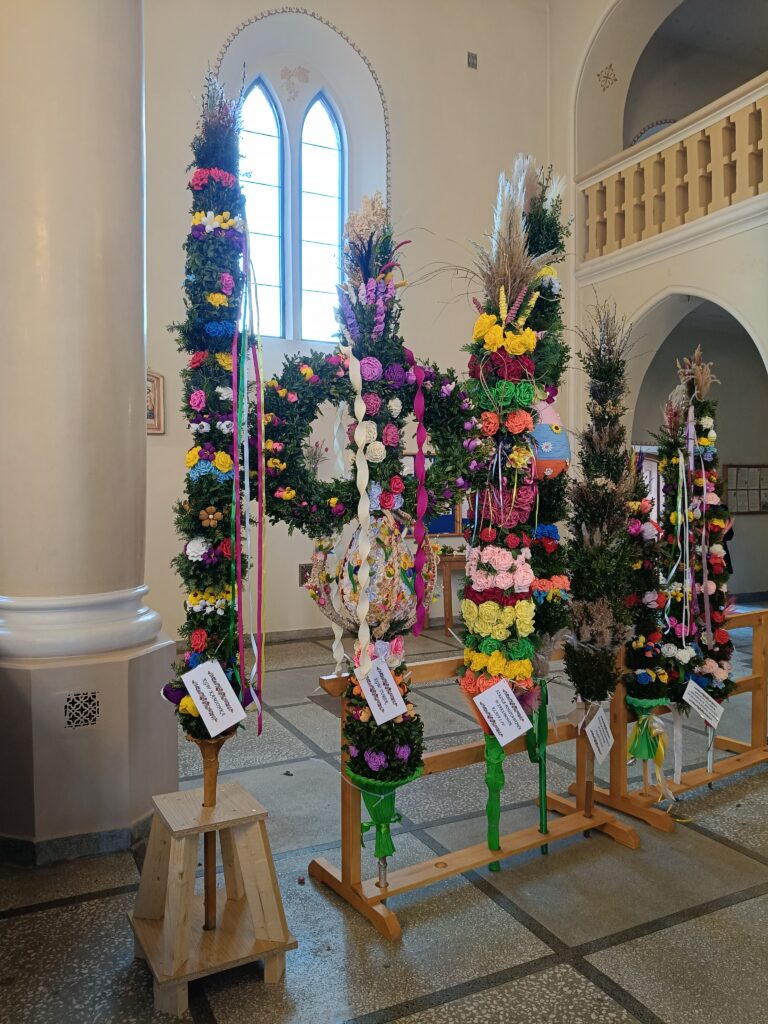 Zdjęcie przedstawia kolorowe palmy wielkanocne stojące w jasnym kościele. Dekoracje mają kwiaty w odcieniach zieleni, żółci, różu i fioletu. Tło to jasna ściana z łukowym oknem.