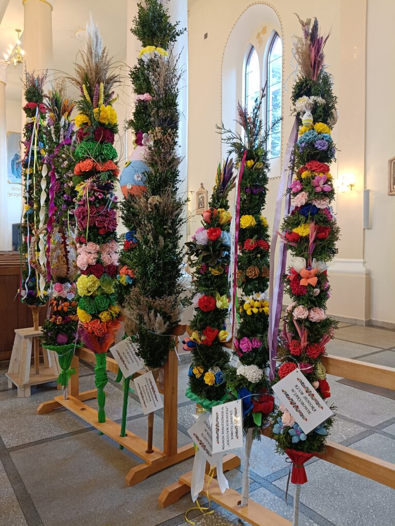 Kolorowe palmy wielkanocne stoją w kościele na drewnianych stojakach. Są ozdobione kwiatami i wstążkami. W tle jasne ściany i okna.